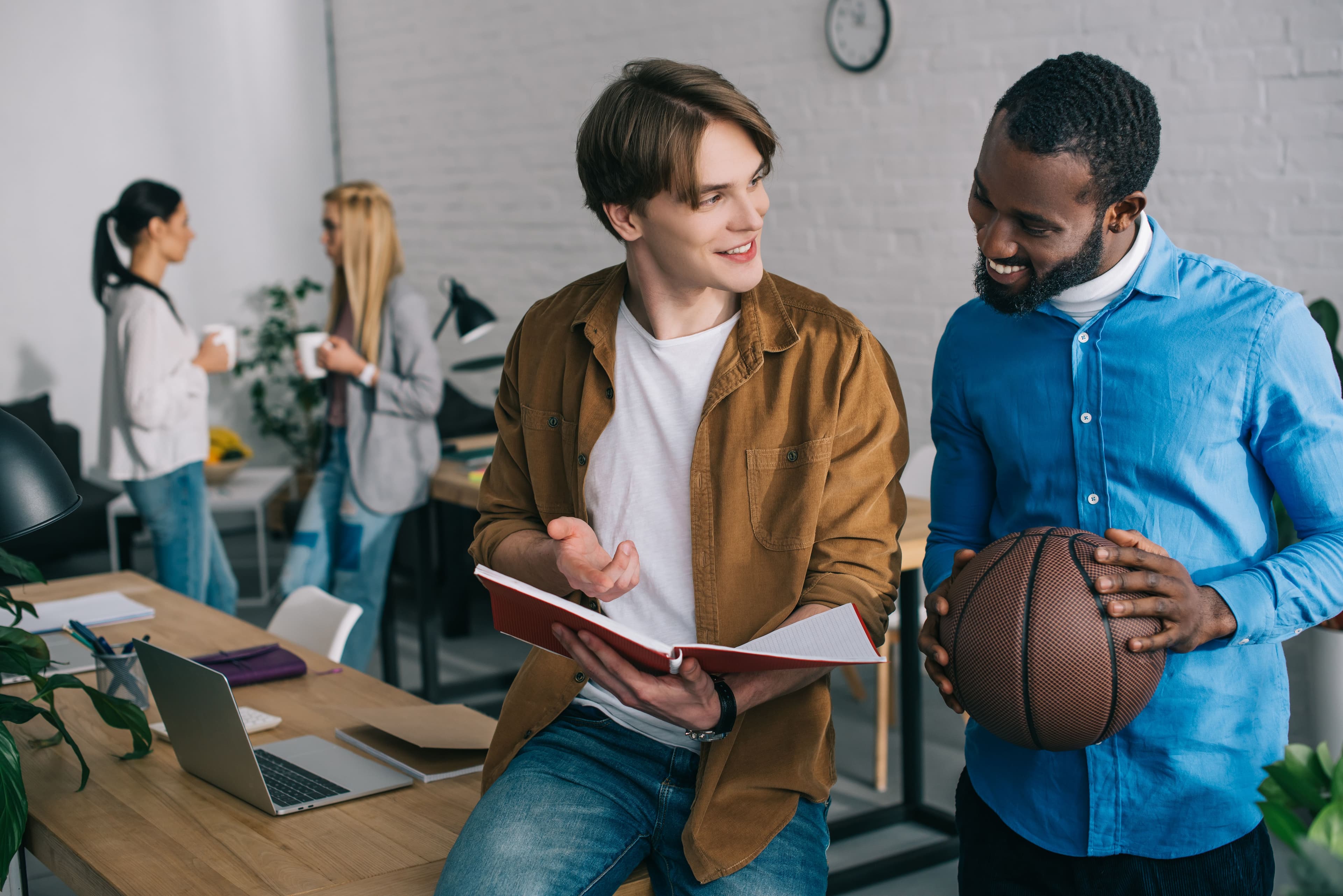 Studenti che discutono durante il corso ITS manager sportivo, con uno di loro che tiene in mano un pallone da basket e altri colleghi sullo sfondo in un ambiente moderno.
